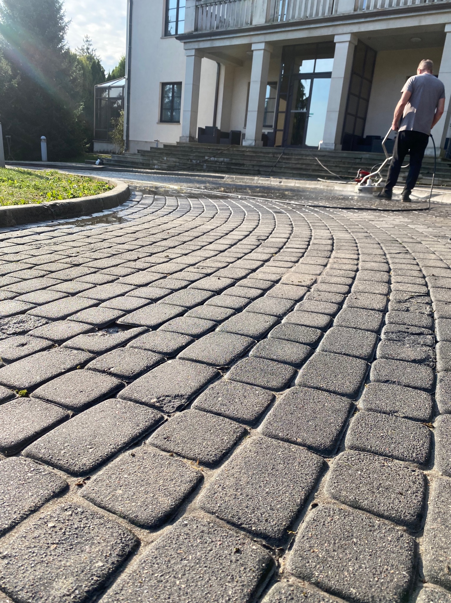 Mężczyzna w trakcie czyszczenia zakrzywionego podjazdu z kostki brukowej za pomocą specjalistycznej maszyny ciśnieniowej; w tle widoczny budynek z kolumnami.
