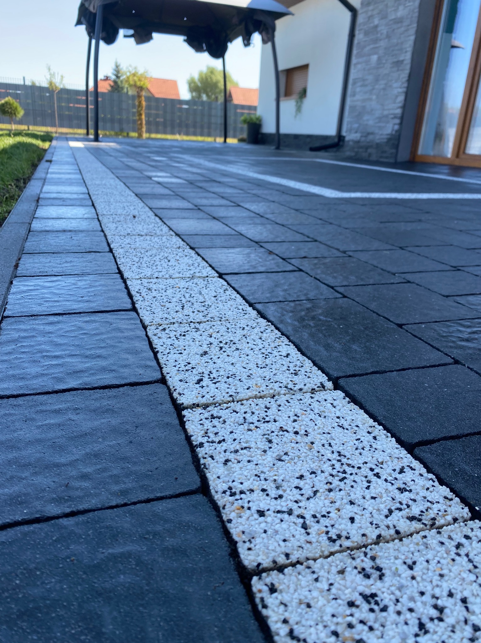 Nowo ułożona kostka brukowa w kolorze grafitowym z białym pasem dekoracyjnym z drobnych kamyczków, widok z niskiej perspektywy na tle domu.
