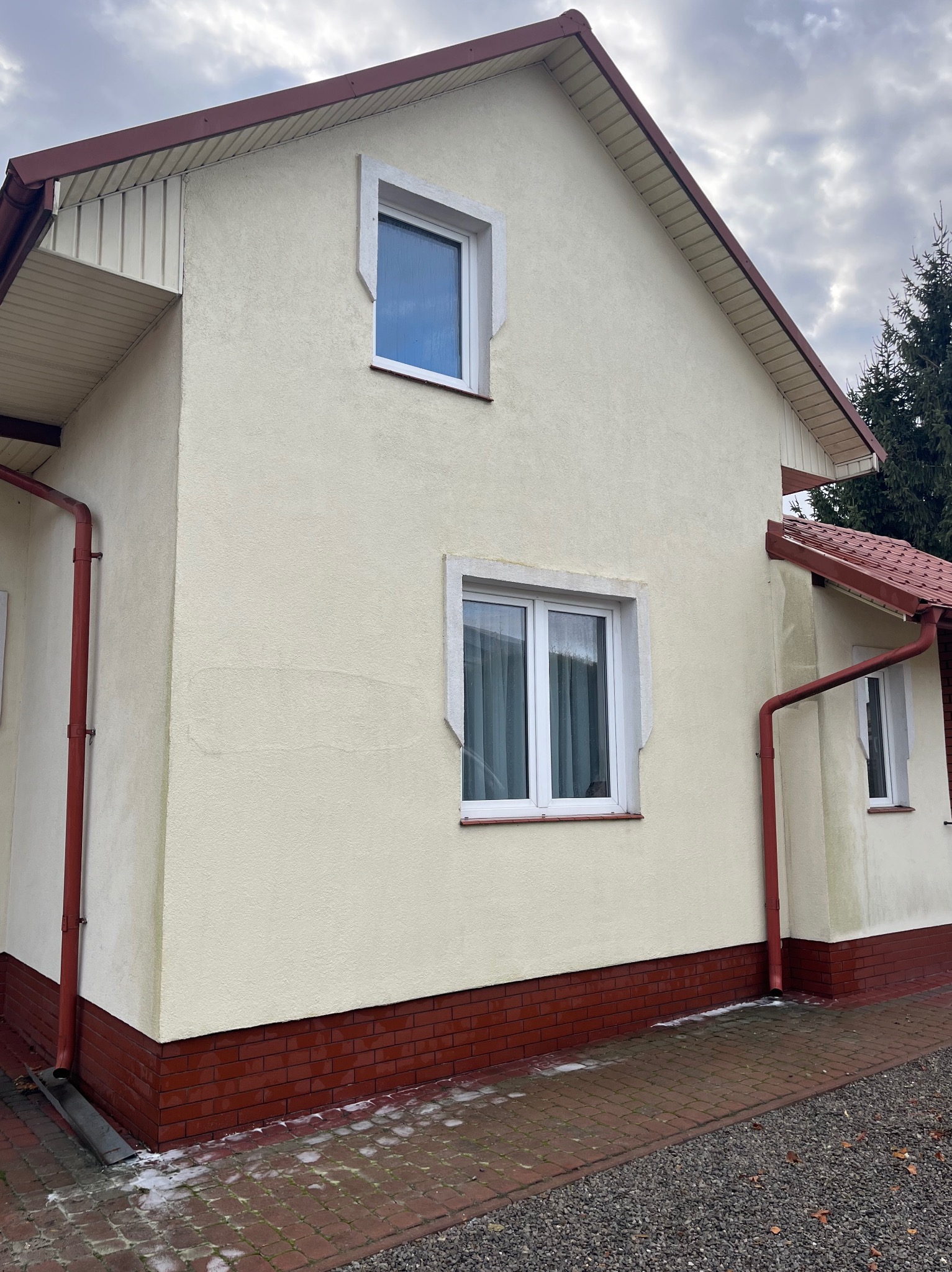 Fragment elewacji domu z widocznymi śladami po czyszczeniu. Jasna elewacja, czerwona ceglana podmurówka, białe okna i czerwone rynny. Częściowo oczyszczona kostka brukowa.