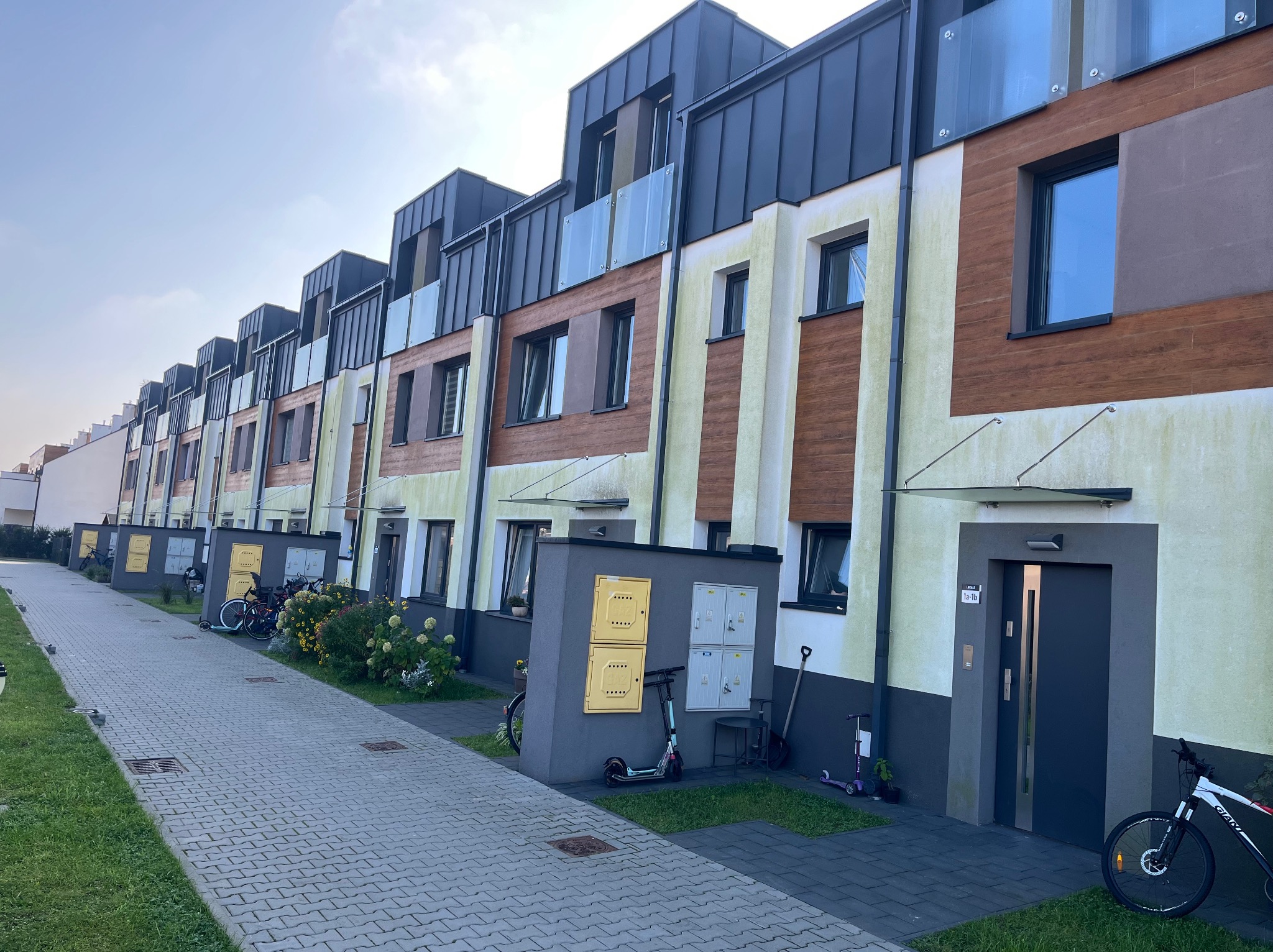 Szeregówka z elewacją w odcieniach beżu i brązu, z widocznymi zabrudzeniami na ścianach. Kostka brukowa przed budynkami, rowery i hulajnogi.