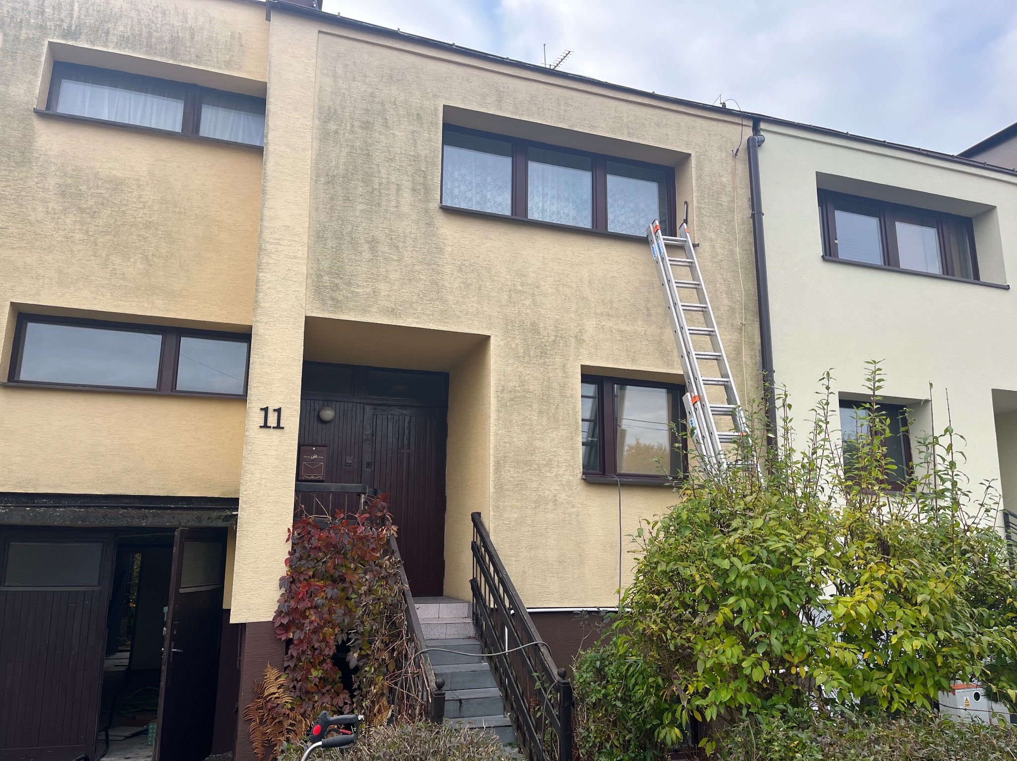 Dwukondygnacyjny dom z elewacją wymagającą odświeżenia, drabina oparta o ścianę, widoczne zabrudzenia i zacieki. Numer domu 11.
