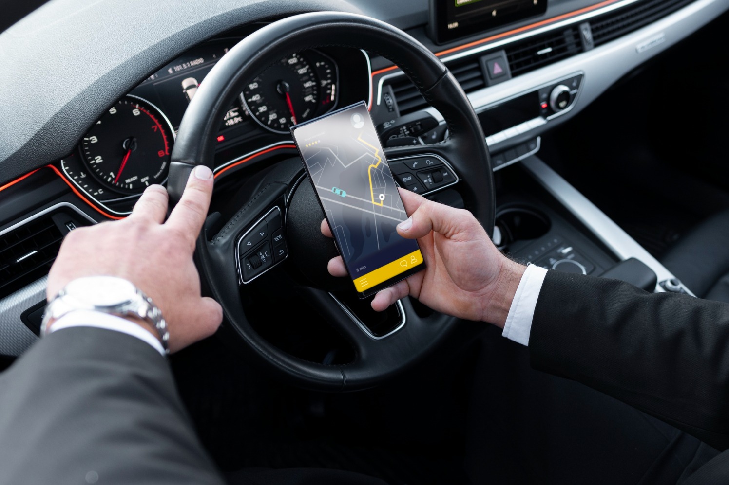 Kierowca w garniturze sprawdza nawigację w telefonie, trzymając kierownicę. Widoczny zegarek na ręce i deska rozdzielcza samochodu z pomarańczowym akcentem.