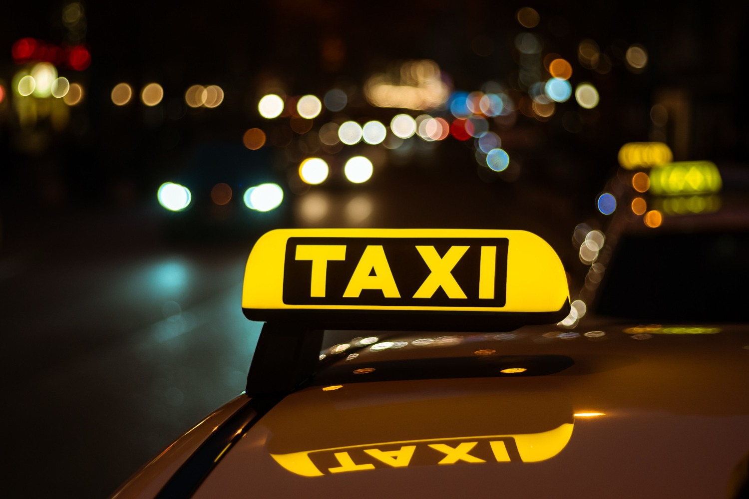 Podświetlony szyld TAXI na dachu samochodu w nocnej scenerii miejskiej z rozmytymi światłami w tle. Widoczne odbicie szyldu na karoserii.