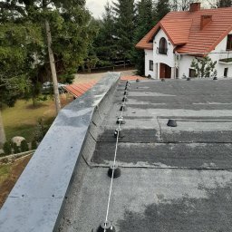 Kamil Duda - Widok z góry na dach pokryty papą z zainstalowaną instalacją odgromową, widoczne uchwyty i przewód, w tle dom z czerwoną dachówką i drzewa.