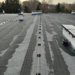 Kamil Duda - Instalacja odgromowa na płaskim dachu pokrytym papą, widoczny pracownik w niebieskiej kurtce i czarne podstawki z przewodami, w tle drzewa i pochmurne niebo.