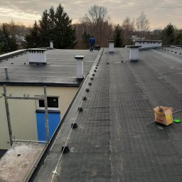 Kamil Duda - Praca na dachu pokrytym papą, widoczne kominy, zamontowana lina odgromowa z uchwytami, pracownik w oddali, karton po materiale na dachu.