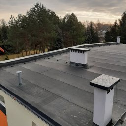 Kamil Duda - Płaski dach pokryty papą termozgrzewalną z trzema kominami wentylacyjnymi na budynku w Skierniewicach. Widoczne mocowania instalacji odgromowej. Pochmurne niebo w tle.