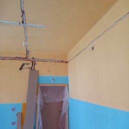 Kamil Duda - Wnętrze pokoju w trakcie remontu; widoczne kable elektryczne prowadzone po ścianach i suficie, częściowo schowane w bruzdach, ściany pomalowane na żółto i niebiesko, drzwi zabezpieczone folią.