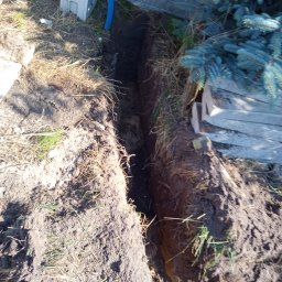 Kamil Duda - Wykop w ziemi z niebieską rurą karbowaną, prawdopodobnie pod kabel elektryczny, widoczne korzenie i trawa na brzegach wykopu, obok biała skrzynka i betonowe panele ogrodzeniowe.