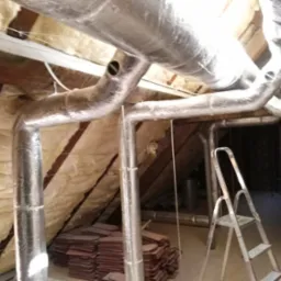 Izolowane rury wentylacyjne na poddaszu, widoczna drabina i stos płytek