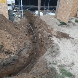 Kamil Duda - Wykop w ziemi z ułożoną rurą kablową, widoczne zabezpieczenia wykopu i materiały budowlane w tle.