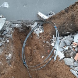 Kamil Duda - Wykonany wykop pod fundament z ułożonymi trzema metalowymi bednarkami połączonymi niebieską taśmą, widoczne gruz i resztki betonu.