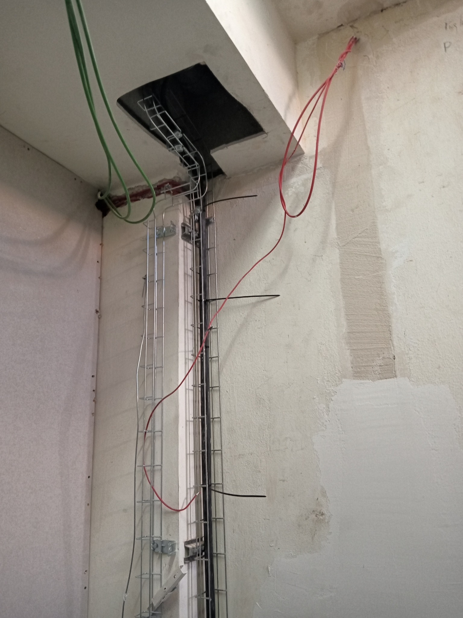 Metalowe korytka kablowe zamontowane na ścianie, z poprowadzonymi przewodami elektrycznymi w kolorach zielonym i czerwonym, widoczne otwory w suficie i ściany w trakcie wykańczania.