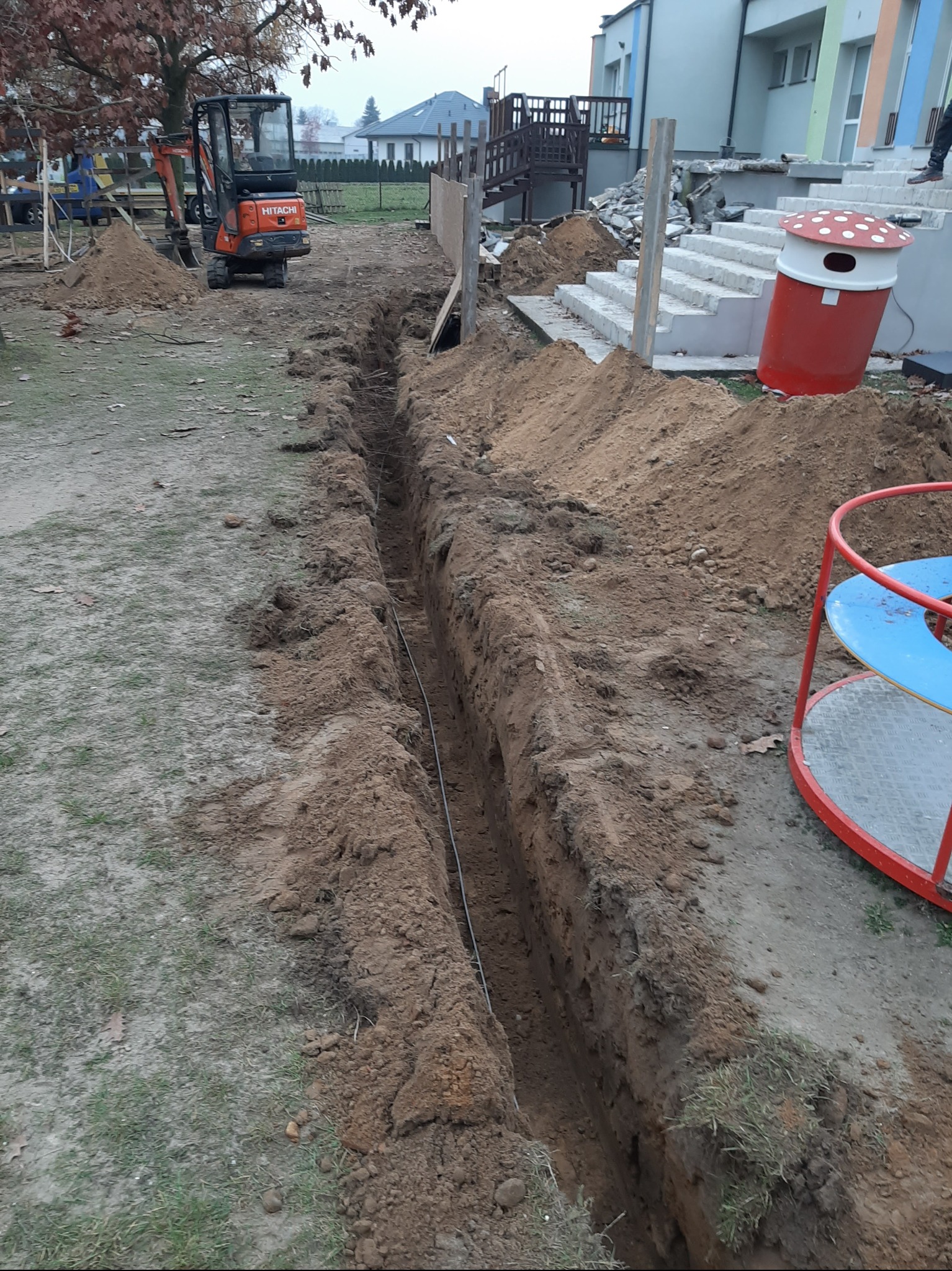 Wykopany rów w ziemi, z widocznym kablem w środku, obok mini koparka Hitachi i elementy placu zabaw w tle.