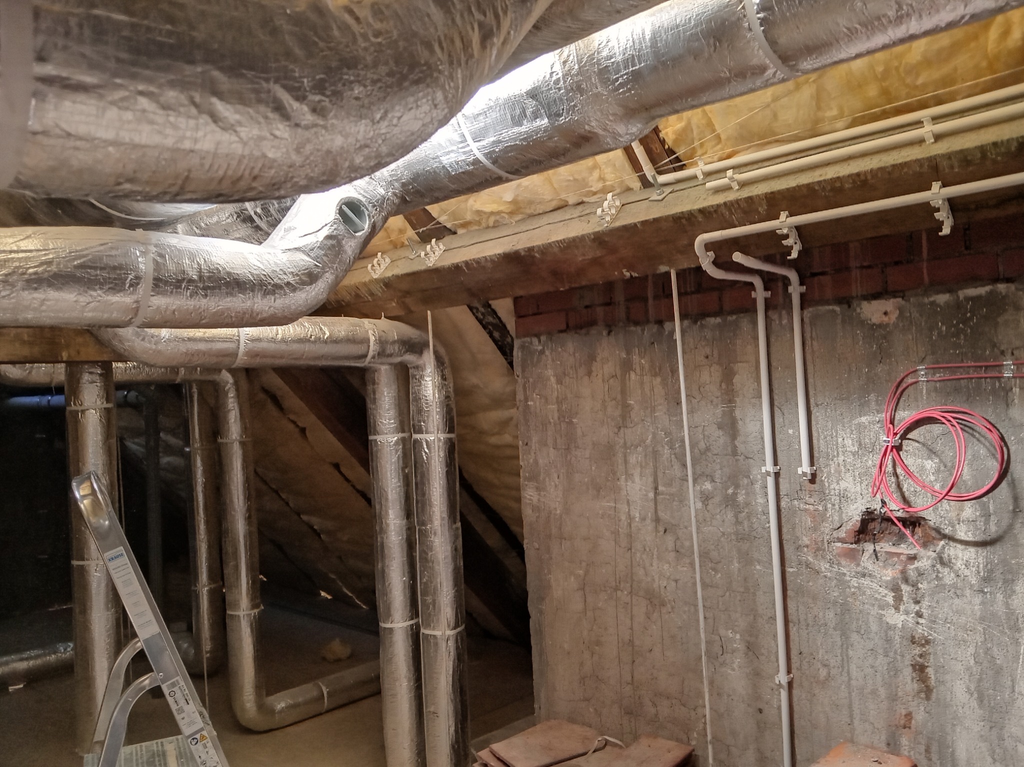 Izolowane rury wentylacyjne pod stropem, widoczne białe rury instalacji wodnej, czerwony kabel elektryczny i drabina aluminiowa.