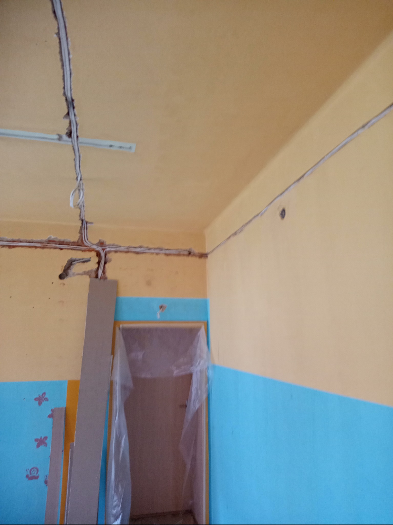 Wnętrze pokoju w trakcie remontu; widoczne kable elektryczne prowadzone po ścianach i suficie, częściowo schowane w bruzdach, ściany pomalowane na żółto i niebiesko, drzwi zabezpieczone folią.