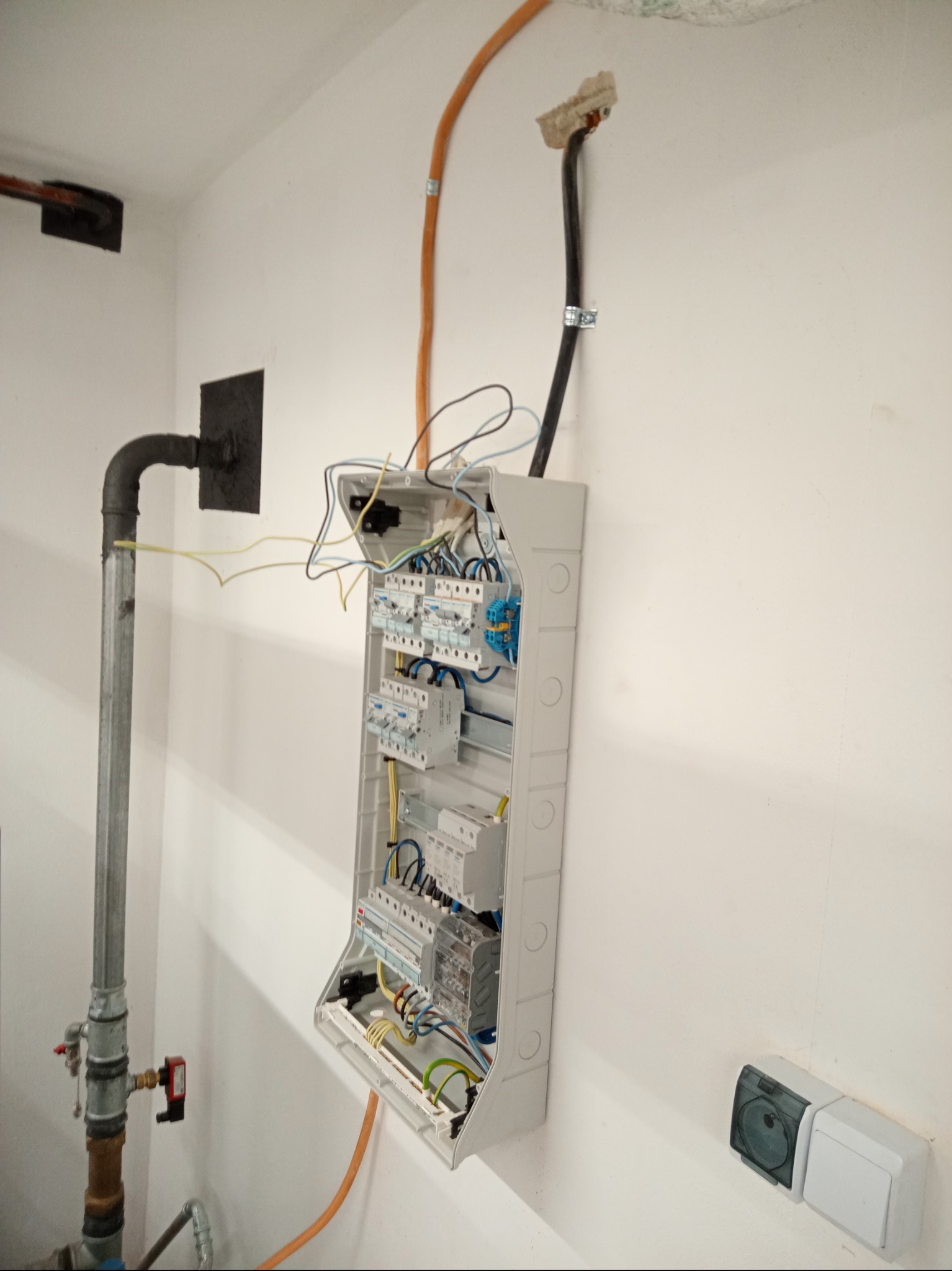 Otwarta skrzynka rozdzielcza z widocznymi bezpiecznikami i przewodami na białej ścianie, obok rury gazowej i gniazdka elektrycznego.