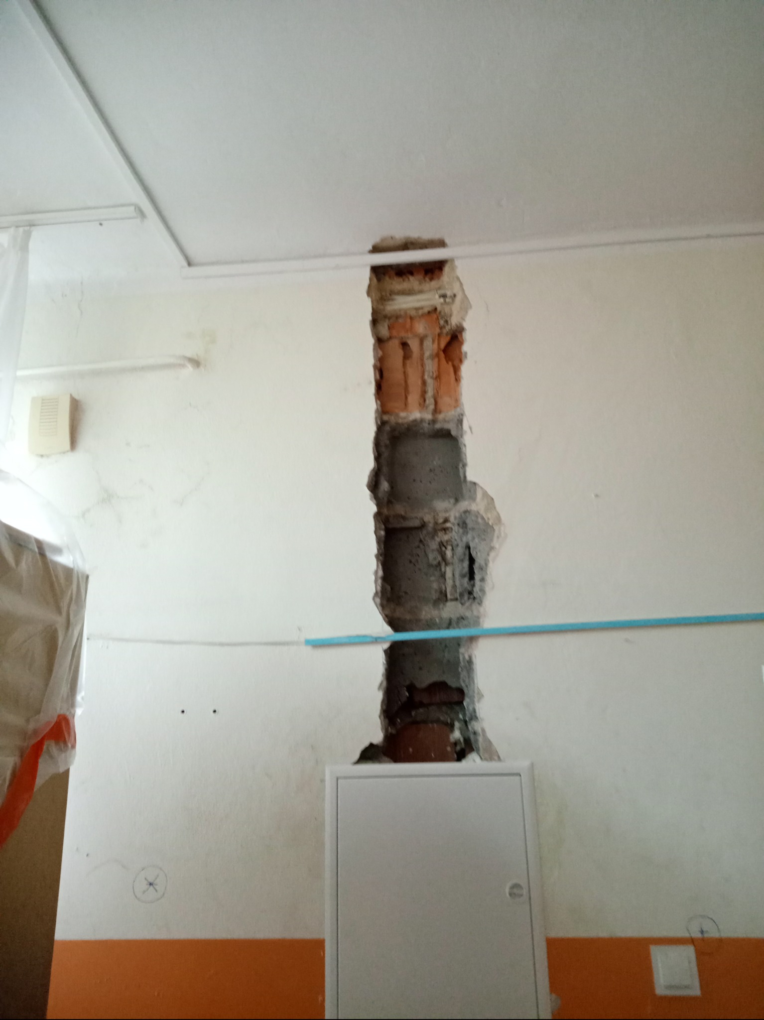 Pionowy otwór w ścianie z widoczną cegłą i betonem, prawdopodobnie pod instalację elektryczną, z białą skrzynką elektryczną na dole i niebieską rurką przechodzącą w poprzek otworu.