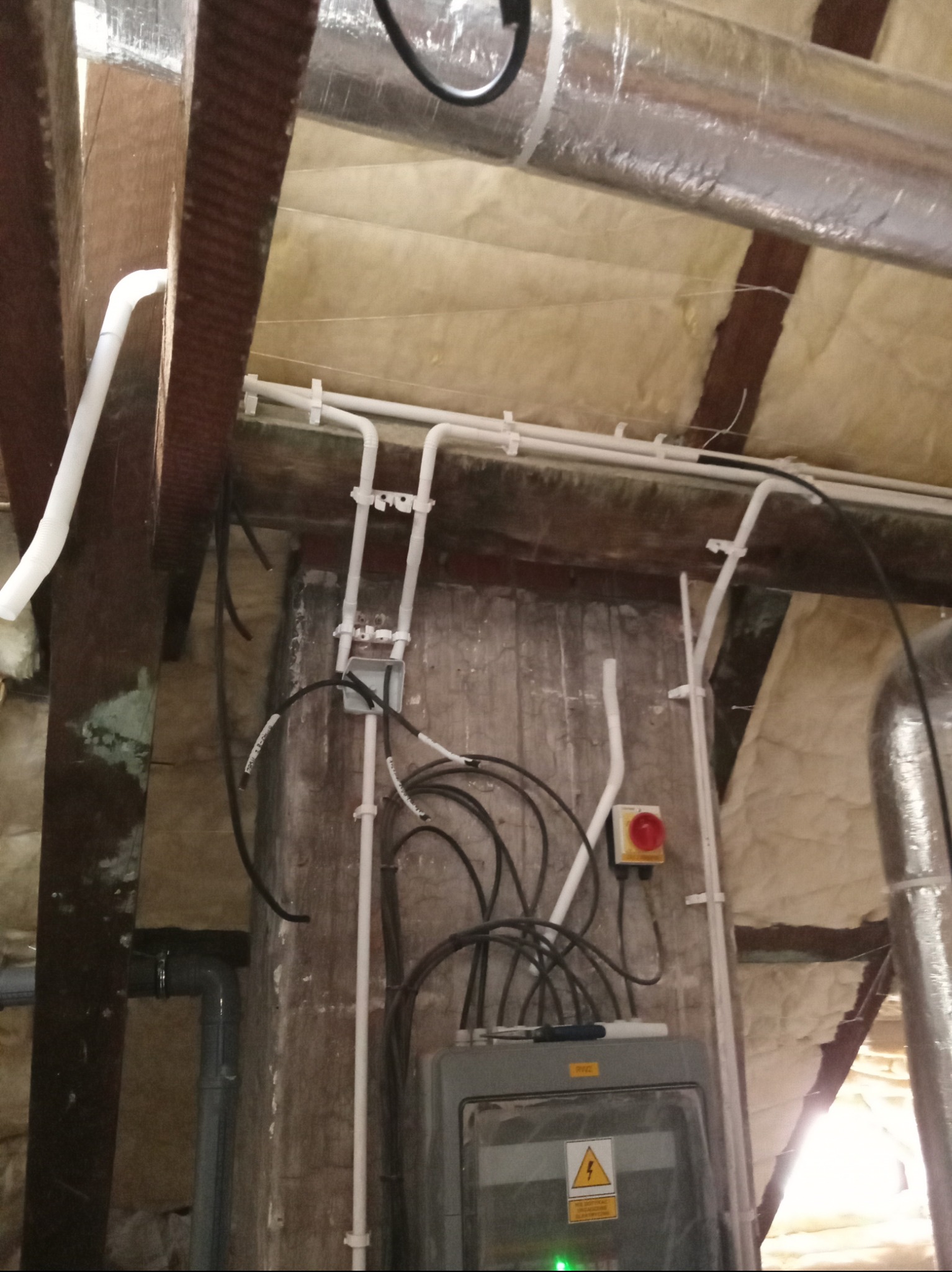 Szara skrzynka elektryczna z żółtym znakiem ostrzegawczym, umieszczona na tle ściany z instalacjami, widoczne przewody elektryczne, rury PCV oraz element konstrukcyjny dachu z izolacją.