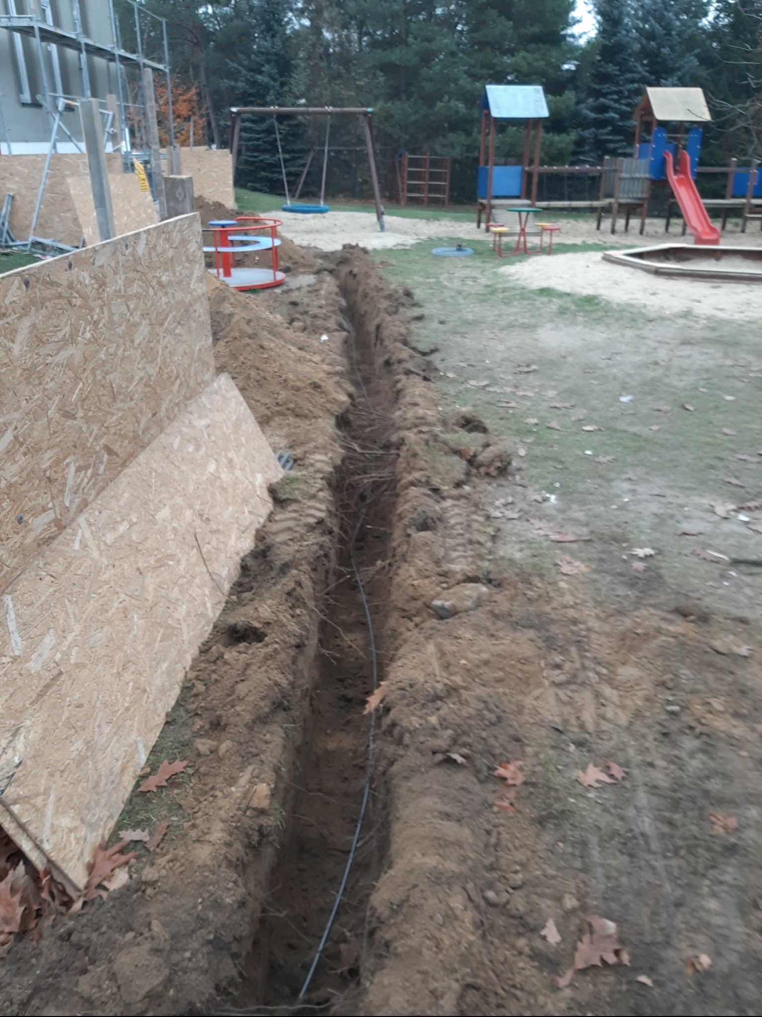 Wkopany przewód w ziemi, widoczny wykop w pobliżu placu zabaw, zabezpieczony płytami OSB.