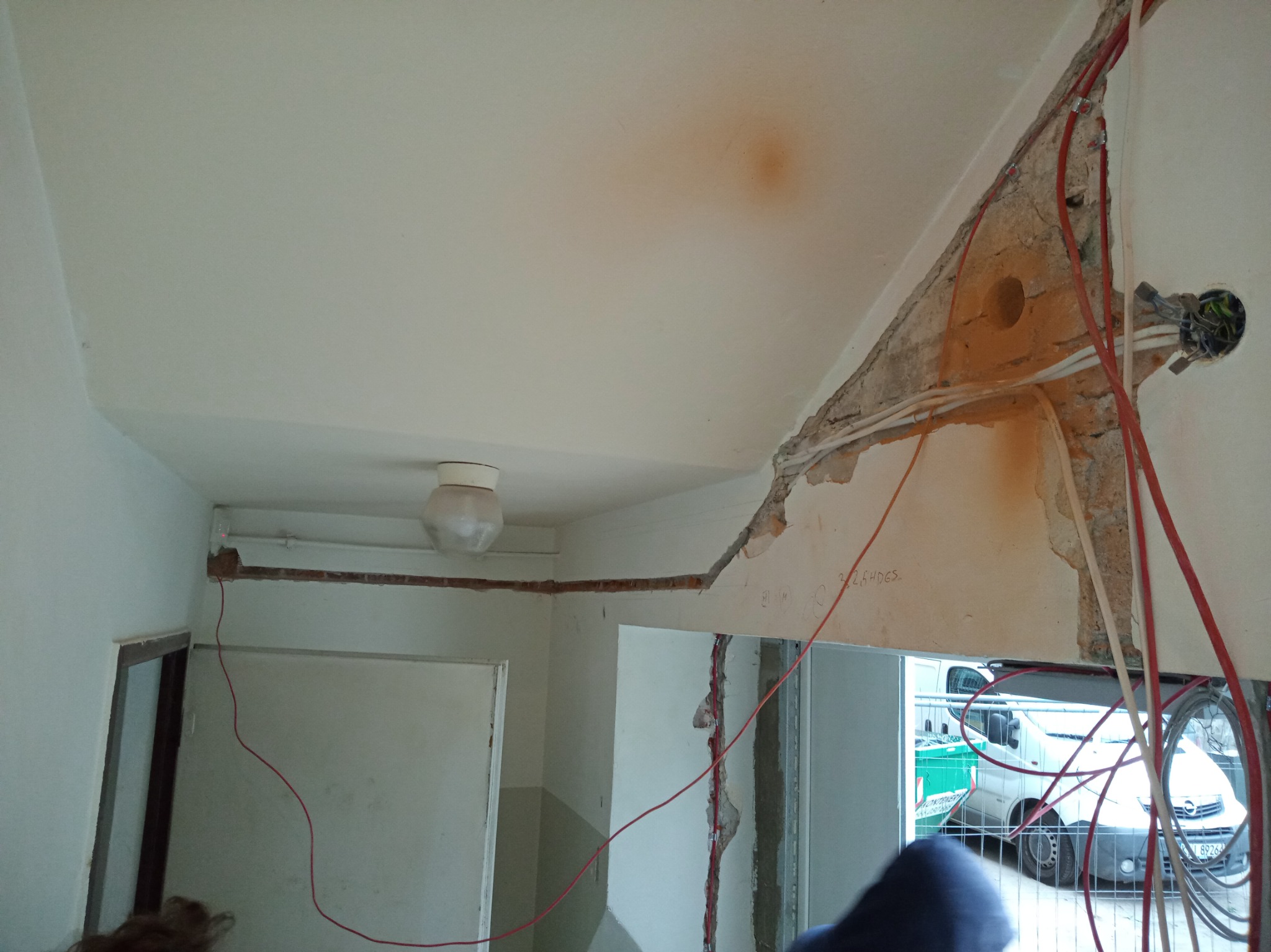 Wnętrze w trakcie remontu z widoczną instalacją elektryczną: przewody w ścianie, puszka elektryczna i wystająca lampa sufitowa. W tle przez otwór w ścianie widoczny samochód dostawczy.