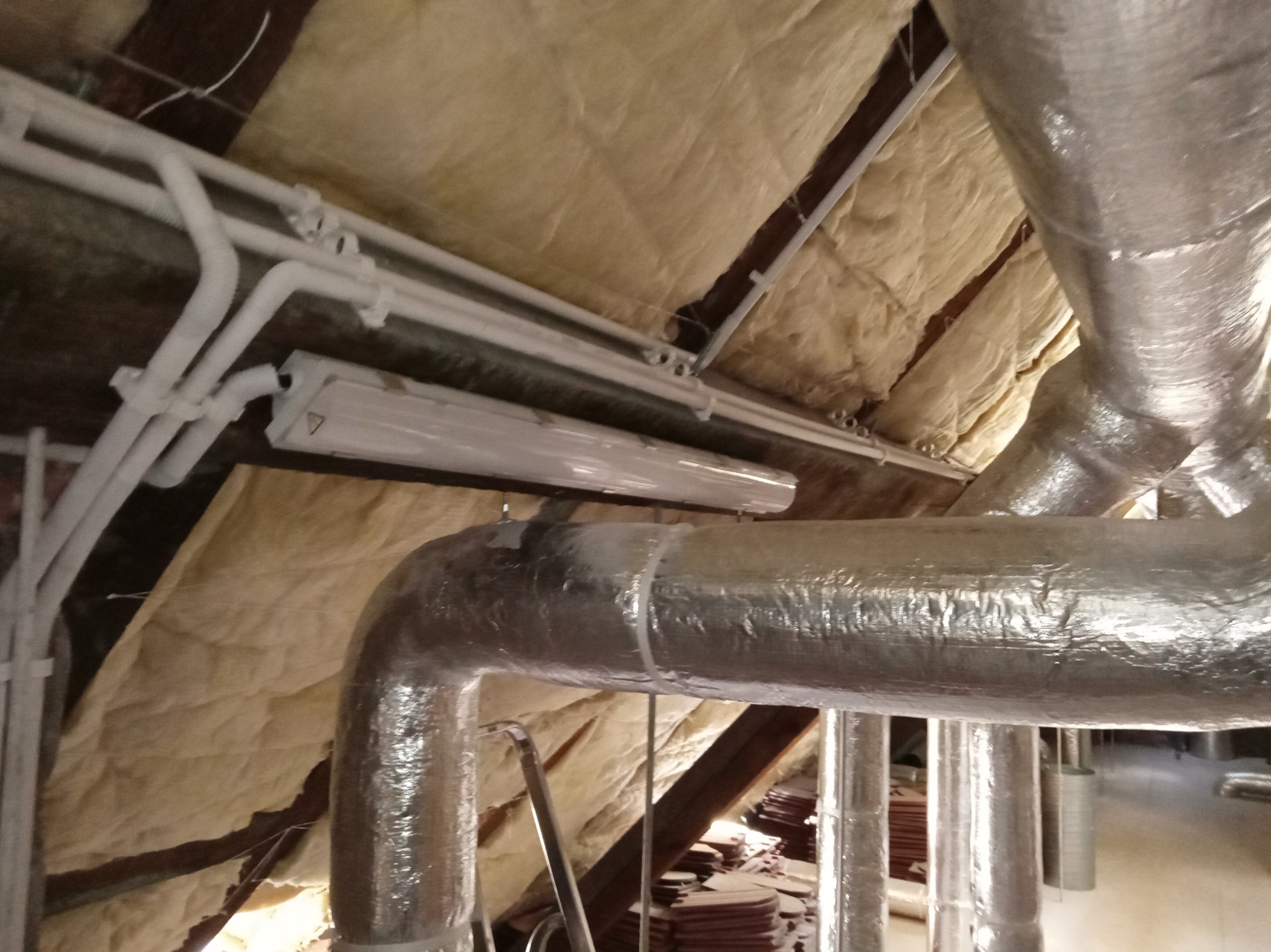 Ocieplone poddasze z widocznymi rurami wentylacyjnymi owiniętymi folią aluminiową oraz białymi rurami z przewodami elektrycznymi i lampą jarzeniową.