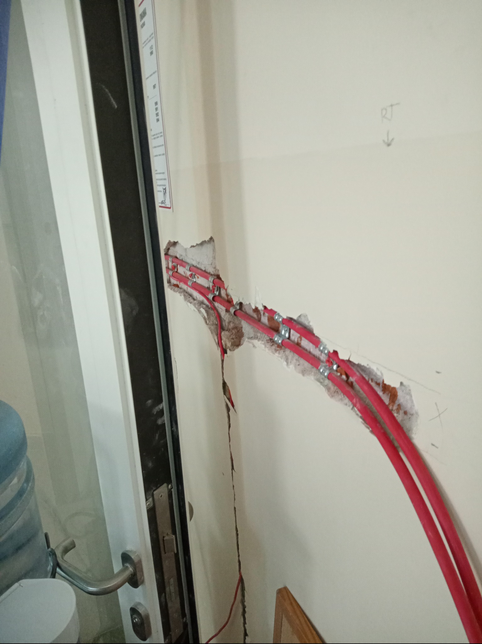 Ściana z wykuciem pod trzy czerwone kable elektryczne, przymocowane metalowymi uchwytami, widoczne ślady wiercenia i notatki na ścianie.