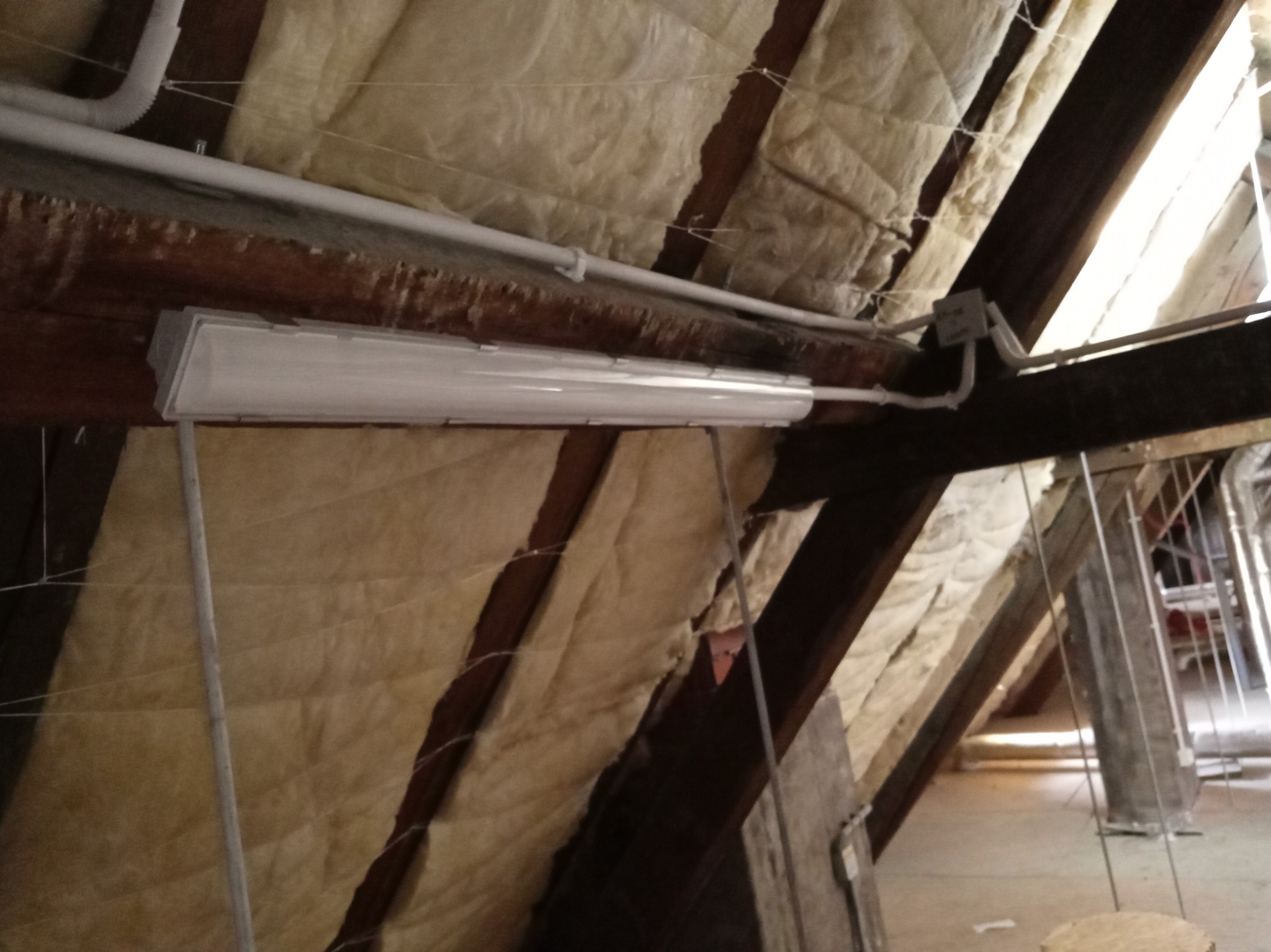 Lampa fluorescencyjna zamontowana pod skosem drewnianej konstrukcji dachu, widoczne przewody elektryczne i izolacja termiczna poddasza.