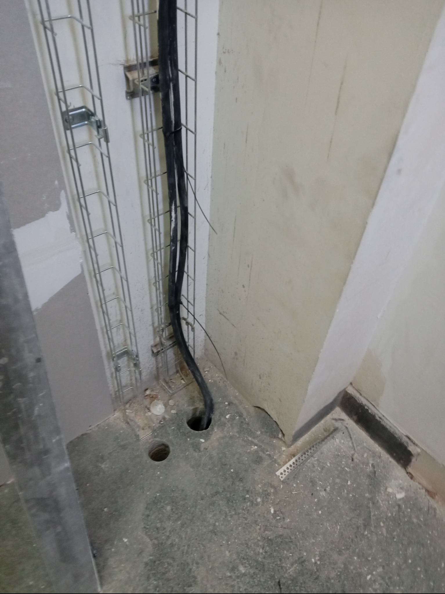 Przewody elektryczne prowadzone w metalowej korycie kablowej, wchodzące w otwór w szarej betonowej podłodze w surowym stanie.