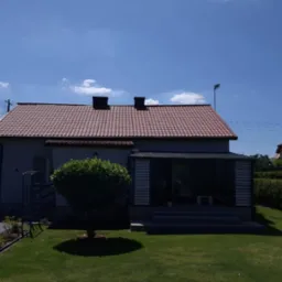 Dom jednorodzinny z czerwonym dachem z dachówki ceramicznej, widok z ogrodu w słoneczny dzień. Widoczne kominy i antena na dachu. Zadbany trawnik i roślinność.