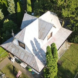 Weronika Simborska - Widok z góry na dom z metalowym dachem, otoczony zielenią. Widoczne drzewa, trawnik i ścieżka. Dach w odcieniach szarości, z widocznymi śladami użytkowania.