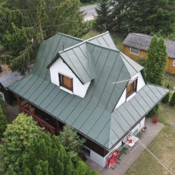 Weronika Simborska - Dom z zielonym, metalowym dachem widziany z góry. Widoczne białe lukarny, antena, drzewa i fragmenty innych budynków w tle. Ujęcie z lotu ptaka.