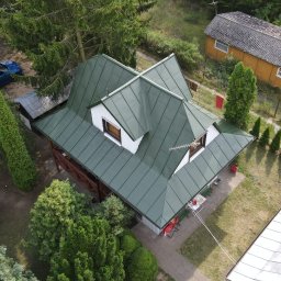 Weronika Simborska - Dom z zielonym dachem z lotu ptaka, otoczony drzewami. Widoczny fragment podwórka, białe okna dachowe i antena. Ujęcie z góry, w tle inne budynki i samochody.