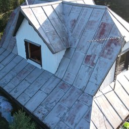 Weronika Simborska - Metalowy dach z widocznymi śladami korozji i łuszczącej się farby, antena na szczycie. Ujęcie z góry, w tle zieleń drzew i fragment niebieskiego samochodu.