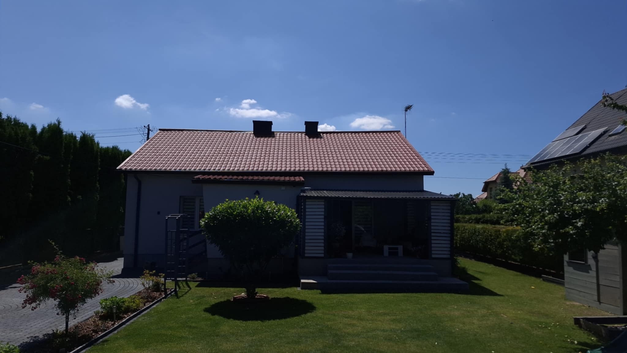 Dom jednorodzinny z czerwonym dachem z dachówki ceramicznej, widok z ogrodu w słoneczny dzień. Widoczne kominy i antena na dachu. Zadbany trawnik i roślinność.