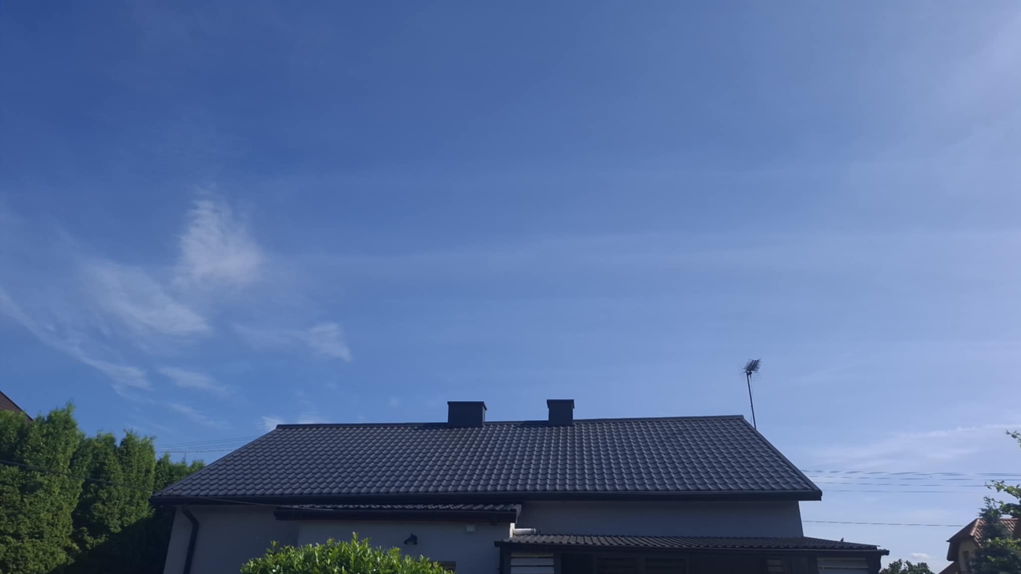 Dom z ciemnoszarym dachem krytym dachówką, widoczny na tle błękitnego nieba z delikatnymi chmurami. Antena na dachu. Częściowo widoczne drzewa i krzewy.