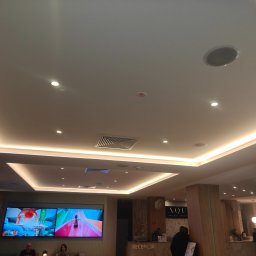 Vitalii Stefaniuk Next - Haus - Sufit podwieszany z oświetleniem LED w recepcji hotelu Aqua Resort. Widoczne kratki wentylacyjne i głośniki sufitowe. Nowoczesny design i jasne kolory.