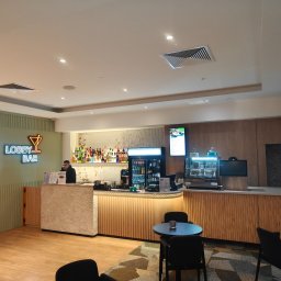 Vitalii Stefaniuk Next - Haus - Nowoczesny bar w hotelowym lobby z neonowym napisem 'LOBBY BAR', drewnianymi panelami i kamienną ladą. Widoczne stoliki i krzesła dla gości.