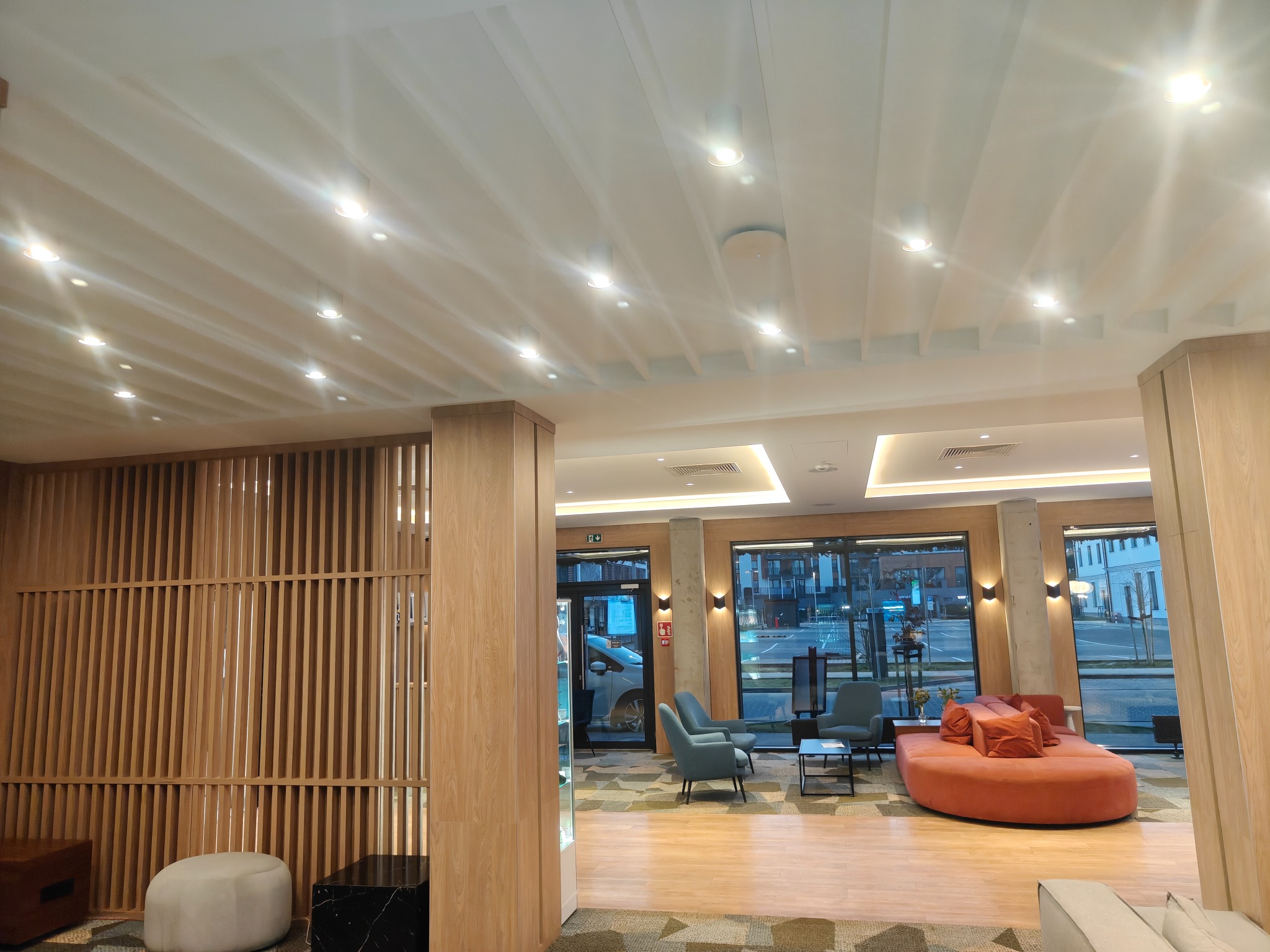 Nowoczesne wykończenie wnętrza biura z drewnianymi panelami, oświetleniem punktowym i designerskimi meblami w odcieniach szarości i pomarańczy. Widok na zewnątrz przez duże okna.