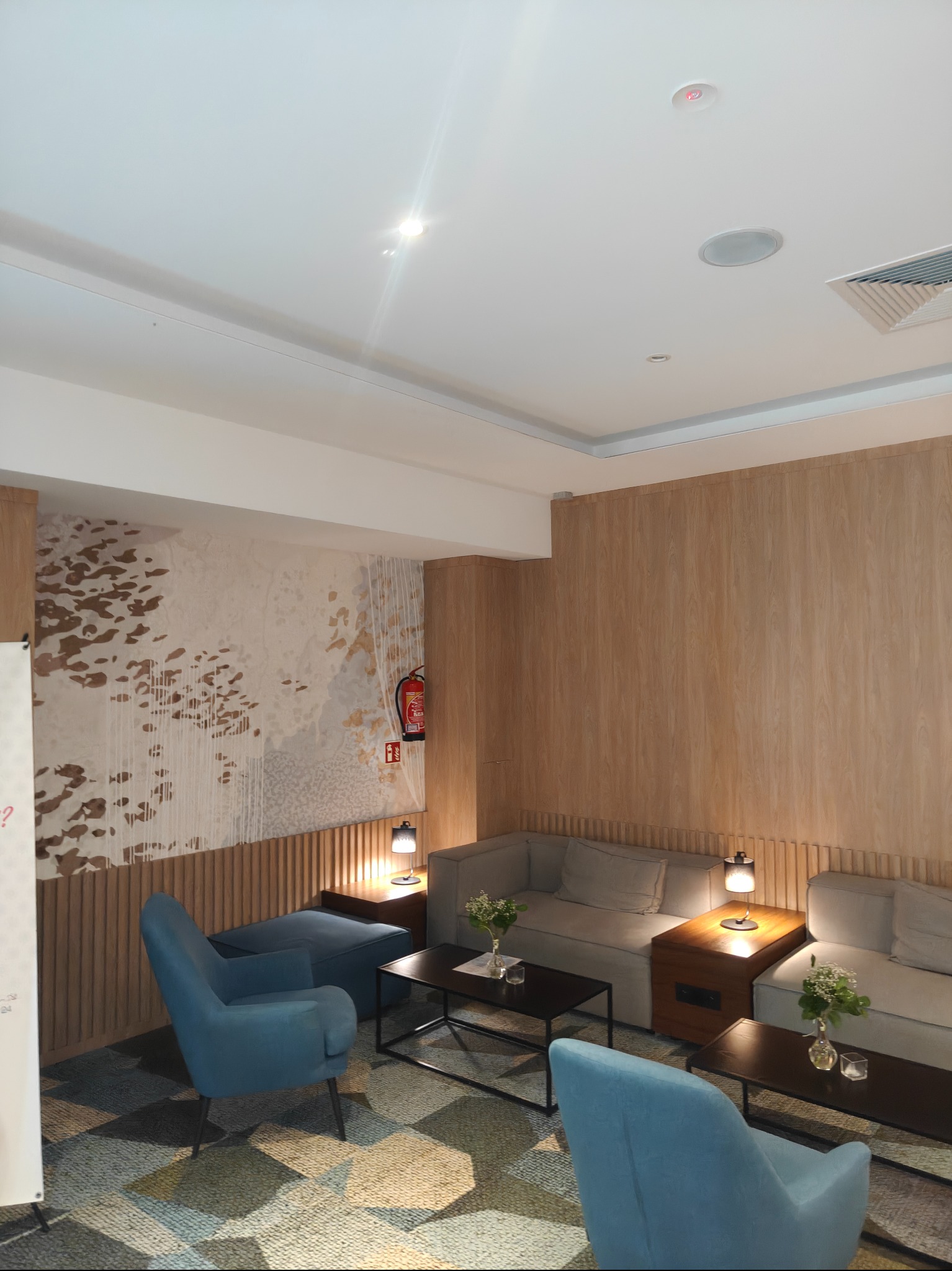 Stylowe wykończenie wnętrza biura: drewniane panele ścienne, abstrakcyjna tapeta, wygodne sofy i fotele w odcieniach szarości i błękitu, stoliki kawowe z kwiatami.