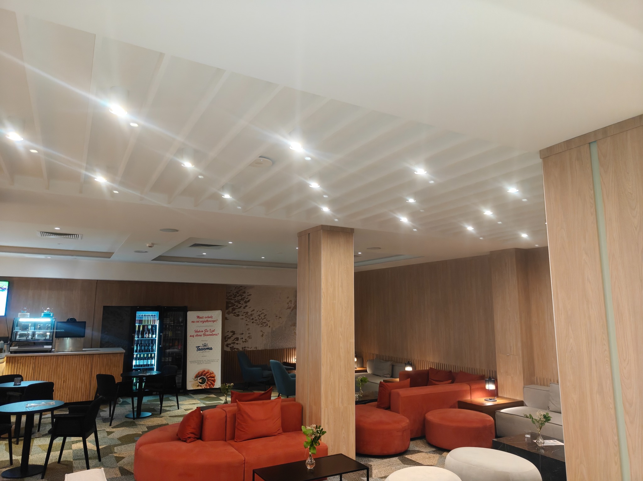 Nowoczesne wnętrze kawiarni z oświetleniem punktowym, drewnianymi panelami ściennymi i pomarańczowymi sofami modułowymi. Widoczny bar i lodówka z napojami.