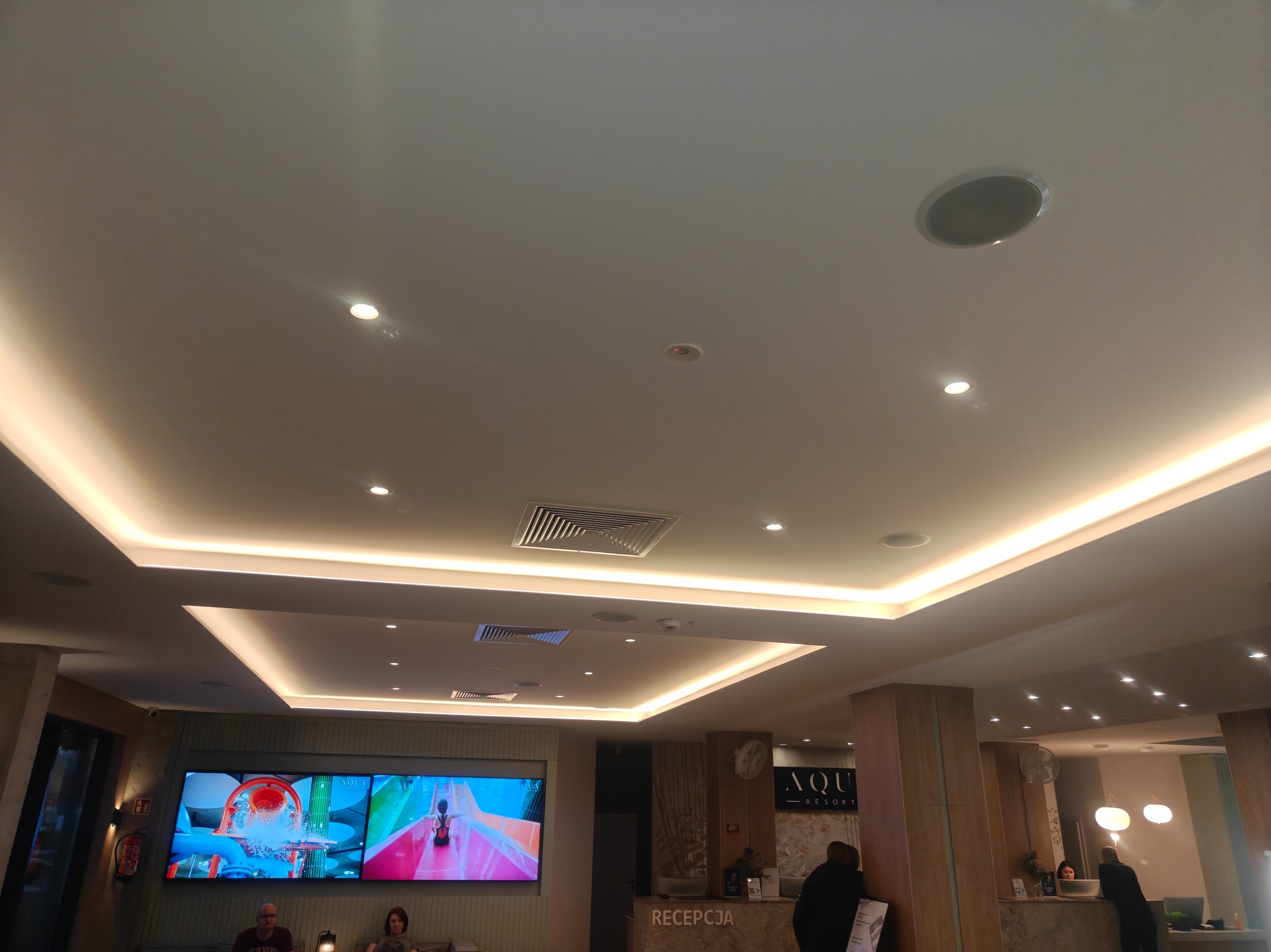 Sufit podwieszany z oświetleniem LED w recepcji hotelu Aqua Resort. Widoczne kratki wentylacyjne i głośniki sufitowe. Nowoczesny design i jasne kolory.