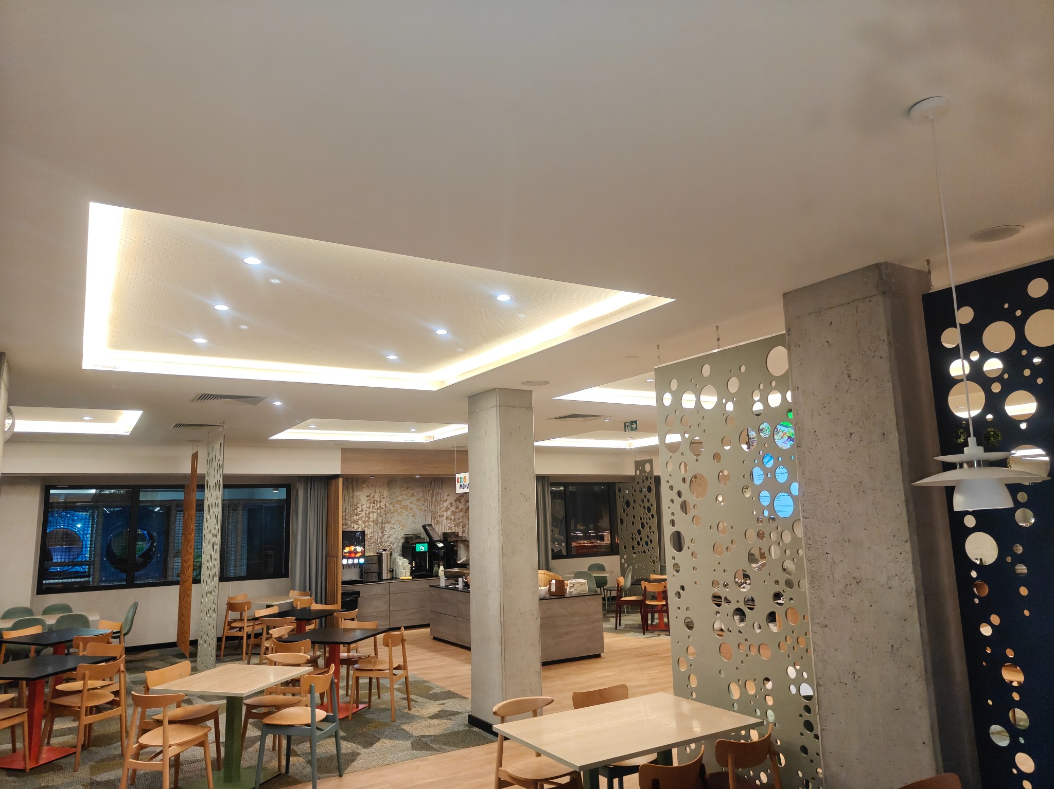 Jasne wnętrze restauracji z drewnianymi krzesłami i stołami, oświetlone sufitami podwieszanymi z wbudowanymi lampami. Dekoracyjne panele z okrągłymi otworami.