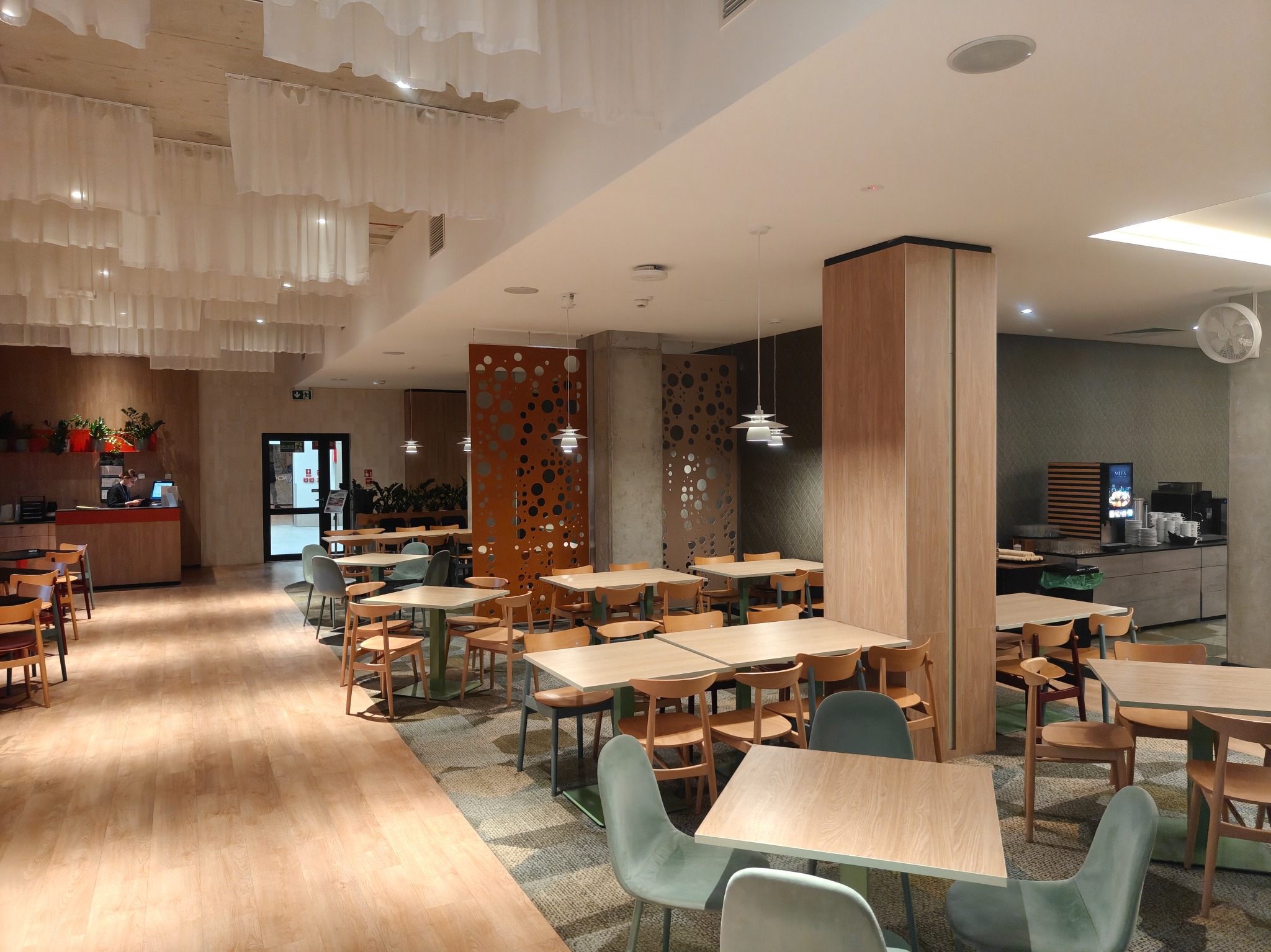 Jasne, przestronne wnętrze restauracji z drewnianą podłogą, stołami i krzesłami. Dekoracyjne, białe tkaniny zwisają z sufitu. Widoczne elementy wystroju i oświetlenia.