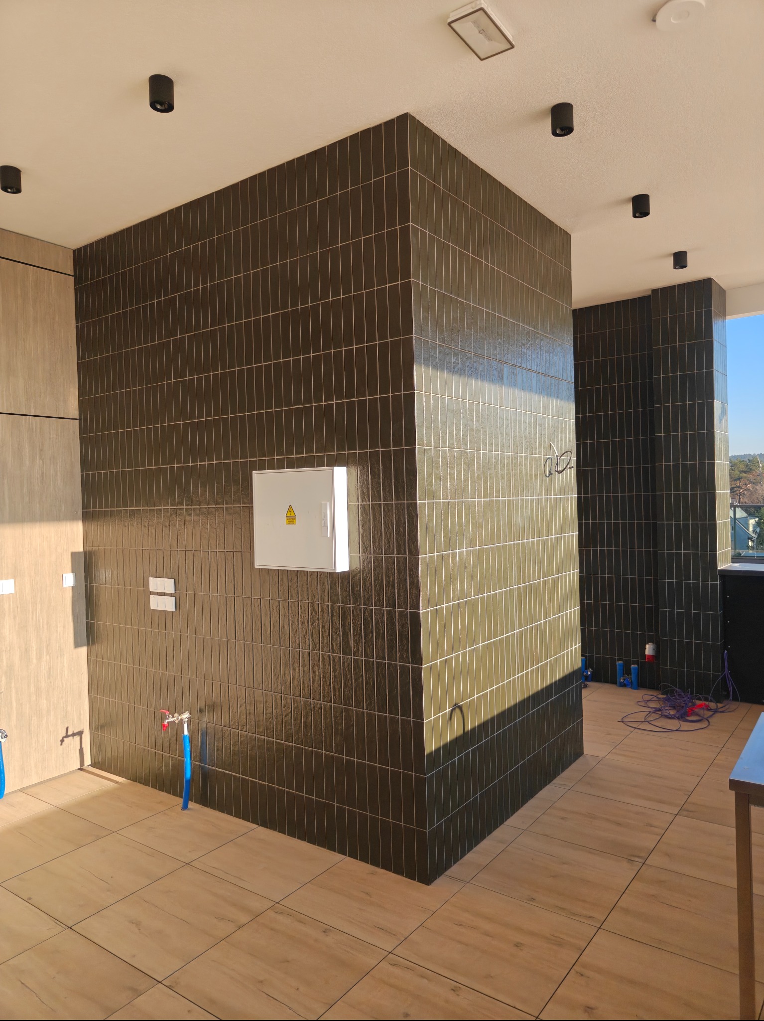 Wnętrze z ciemnymi, pionowo ułożonymi płytkami na ścianach i drewnopodobną podłogą. Widoczne instalacje, skrzynka elektryczna i punktowe oświetlenie sufitowe.