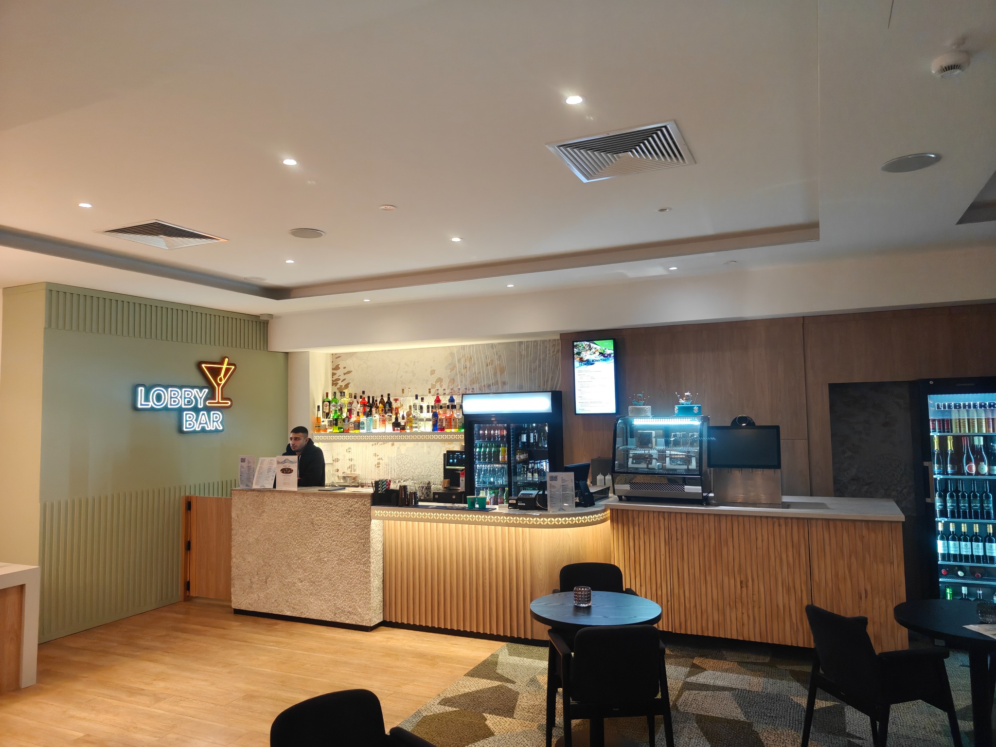 Nowoczesny bar w hotelowym lobby z neonowym napisem 'LOBBY BAR', drewnianymi panelami i kamienną ladą. Widoczne stoliki i krzesła dla gości.