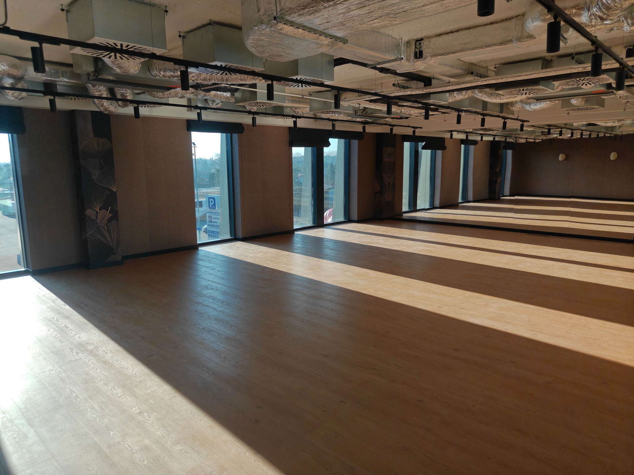 Jasne panele podłogowe w przestronnym biurze z dużymi oknami i widoczną instalacją wentylacyjną pod sufitem. Minimalistyczny design, naturalne światło.