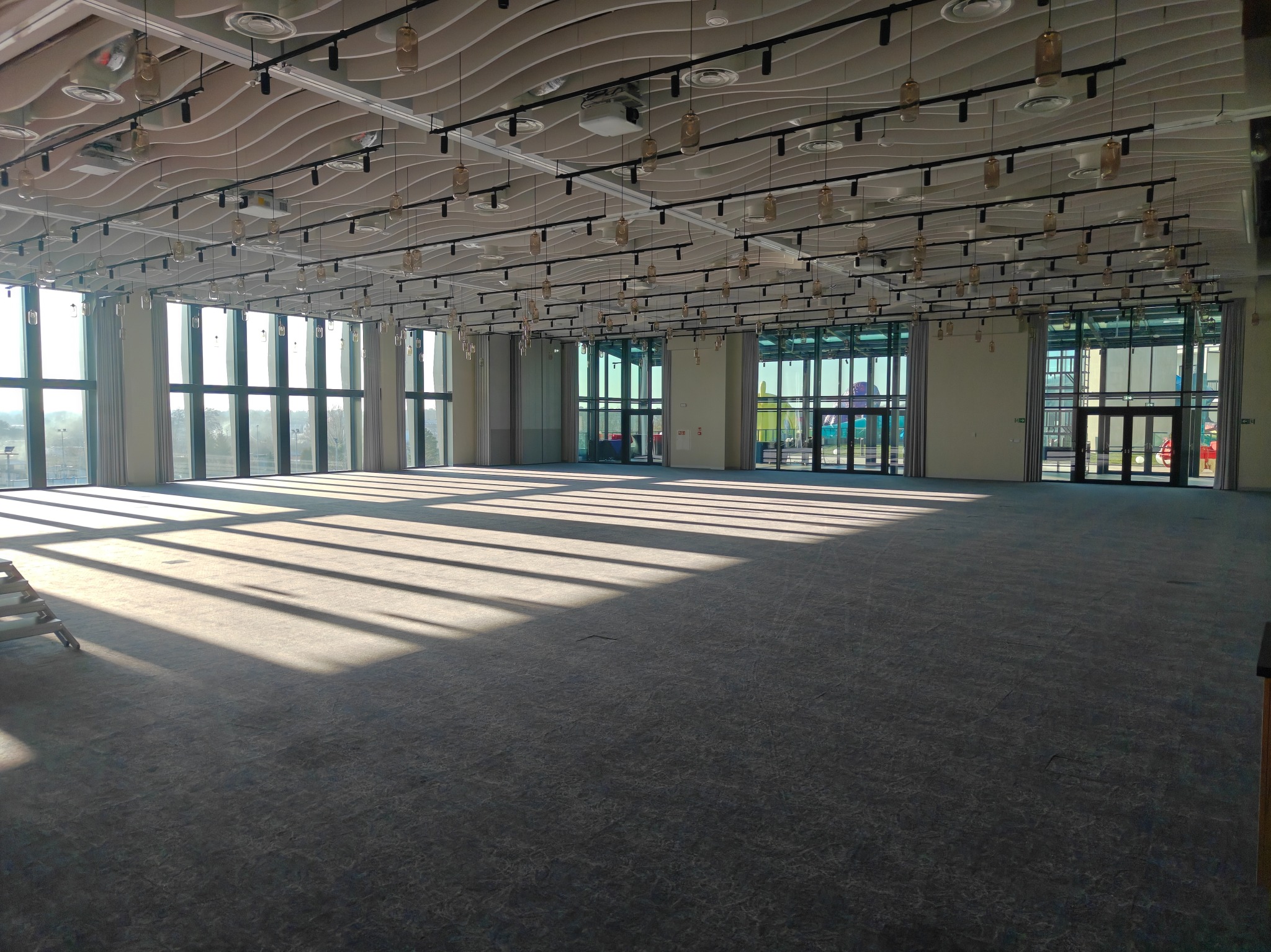 Przestronne, jasne wnętrze sali konferencyjnej z falistym sufitem i rzędami lamp. Promienie słoneczne wpadają przez okna, tworząc geometryczne wzory na wykładzinie.