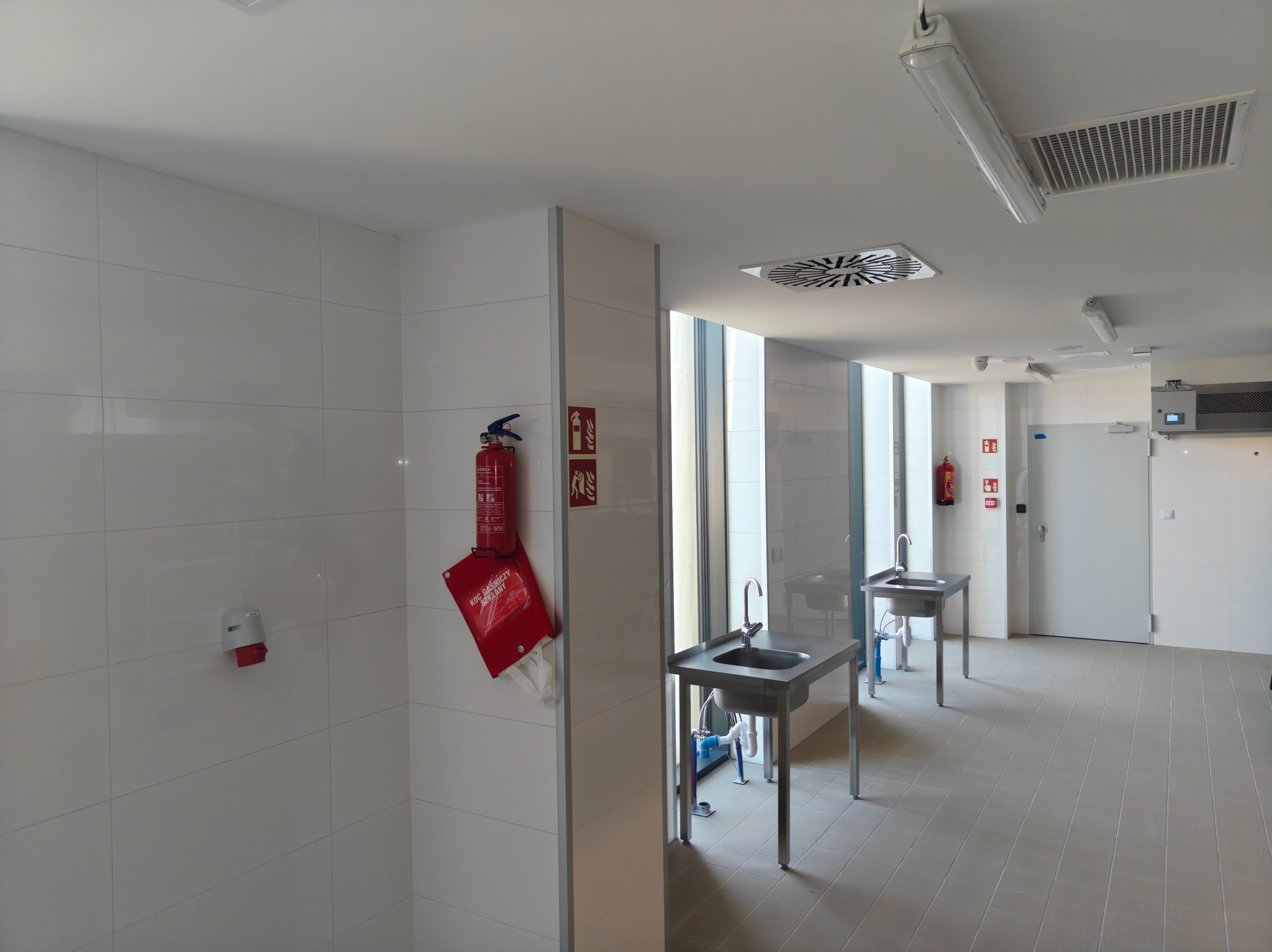 Białe płytki na ścianach i szara podłoga w pomieszczeniu z umywalkami na metalowych stelażach. Widoczne gaśnice i wentylacja sufitowa.