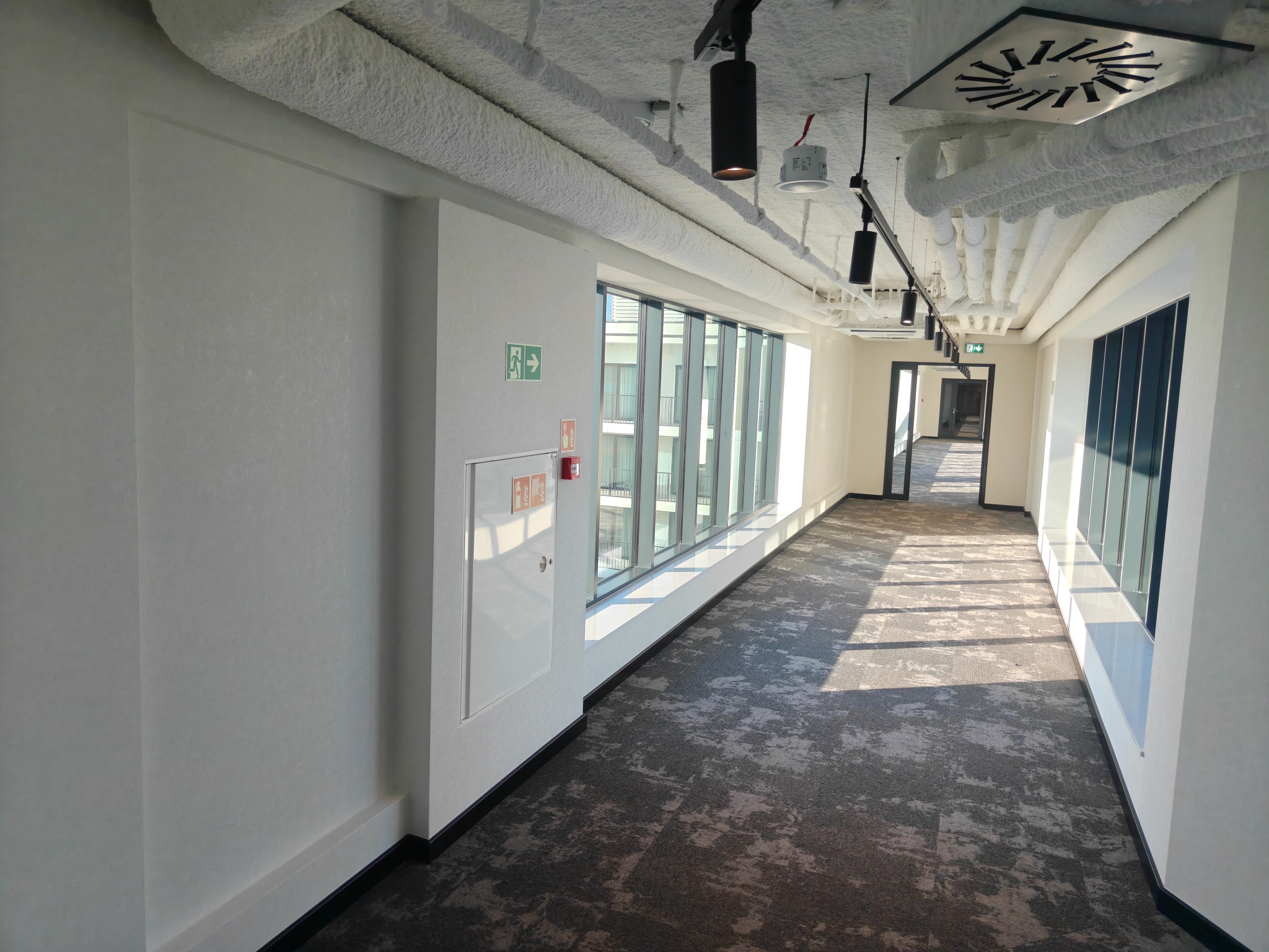 Korytarz biurowy z wykładziną dywanową, białymi ścianami i oknami. Sufit z instalacjami pokrytymi pianką izolacyjną. Widoczny znak ewakuacyjny i hydrant.